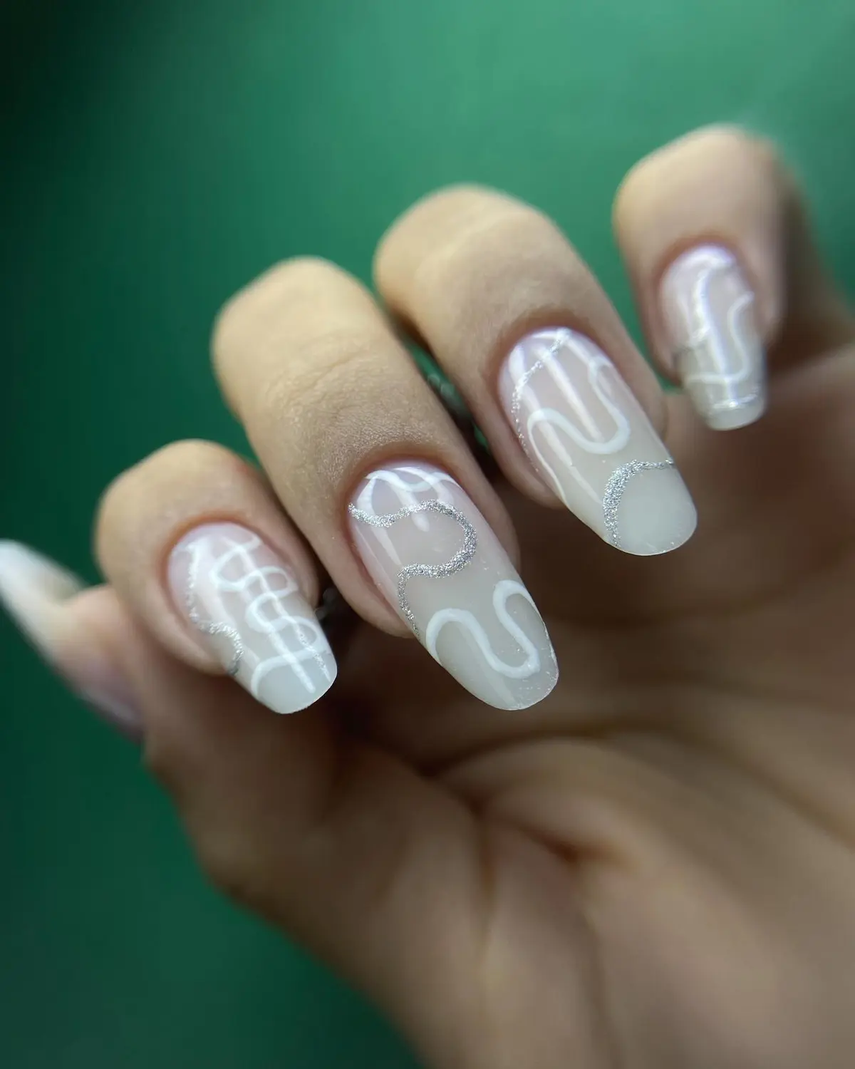 Unhas amendoadas em nude leitoso com desenhos em linhas brancas e prateadas.